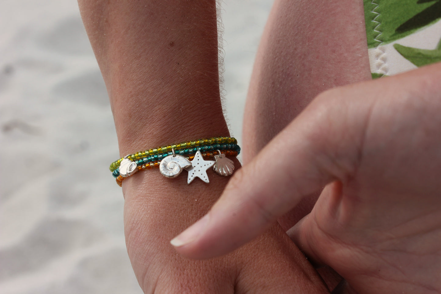 Bracelets en perles de rocailles colorées avec des charms en argent 925 inspirés du bord de mer, avec étoile de mer, coquillage et ammonite, portés au poignet sur une plage ensoleillée.
