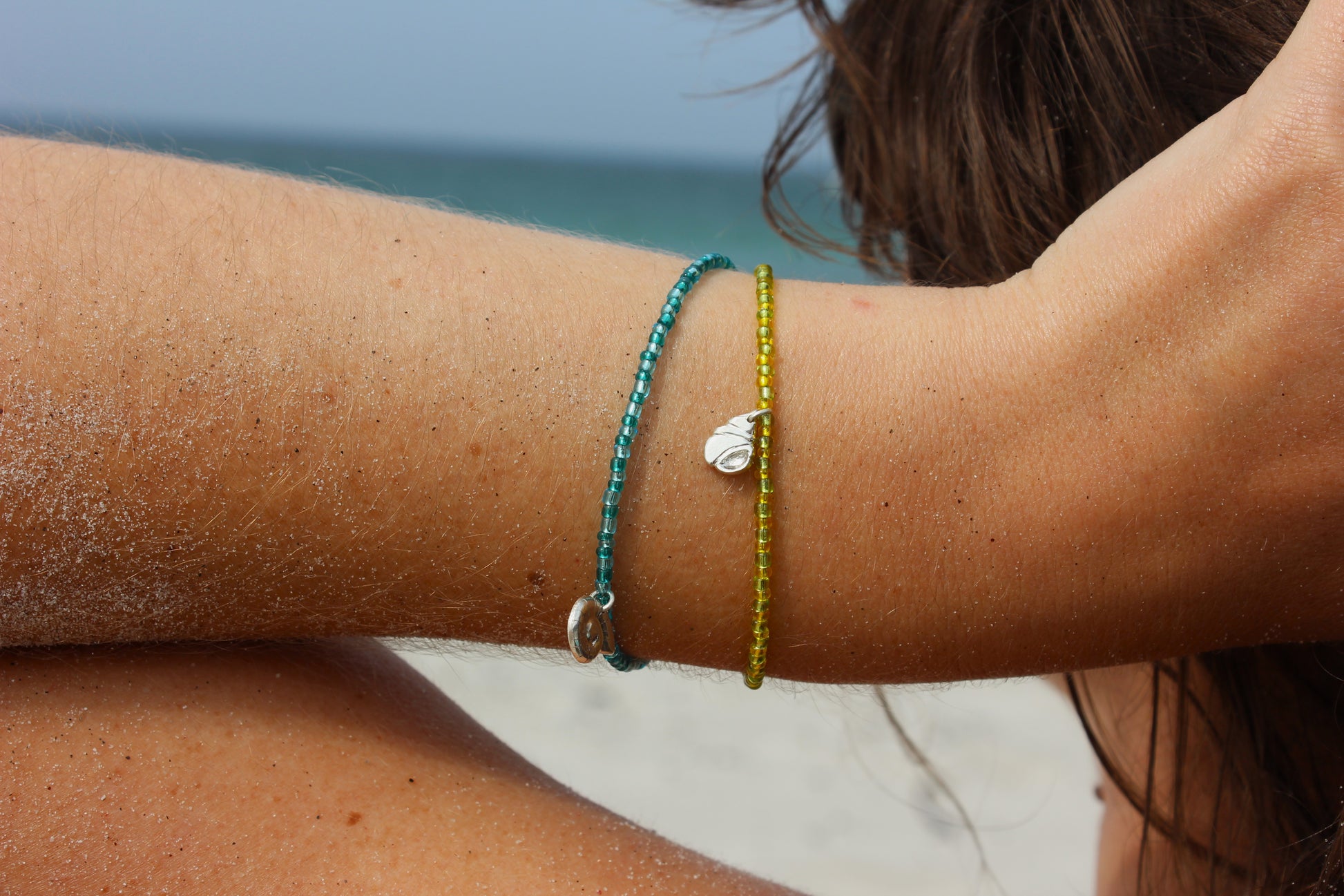 Bracelets en perles de rocailles colorées avec des charms en argent 925 inspirés du bord de mer, avec étoile de mer, coquillage et ammonite, portés au poignet sur une plage ensoleillée.
