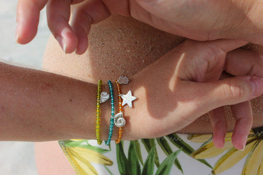 Bracelets en perles de rocailles colorées avec des charms en argent 925 inspirés du bord de mer, avec étoile de mer, coquillage et ammonite, portés au poignet sur une plage ensoleillée.