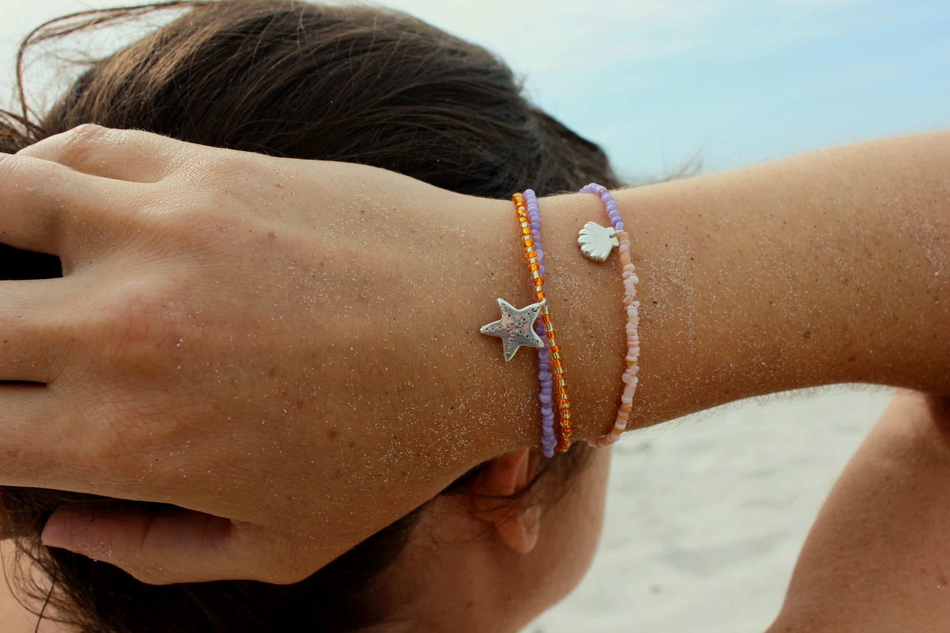 Bracelets en perles de rocailles colorées avec des charms en argent 925 inspirés du bord de mer, avec étoile de mer, coquillage et ammonite, portés au poignet sur une plage ensoleillée.
