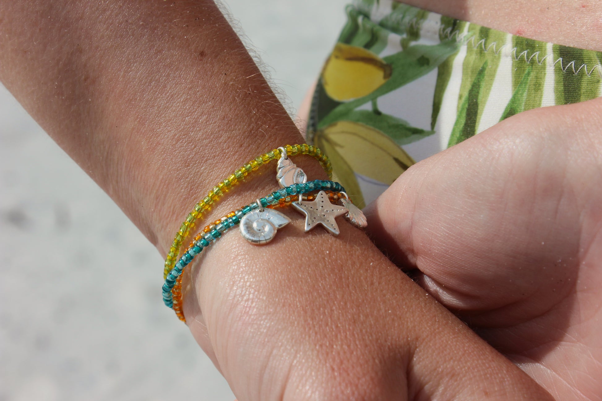 Bracelets en perles de rocailles colorées avec des charms en argent 925 inspirés du bord de mer, avec étoile de mer, coquillage et ammonite, portés au poignet sur une plage ensoleillée.